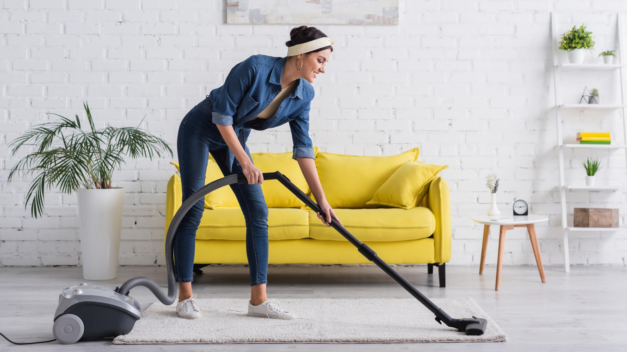 A woman vacuums a light carpet in a modern living room with a yellow sofa and a plant, wearing casual denim clothes and a headband.